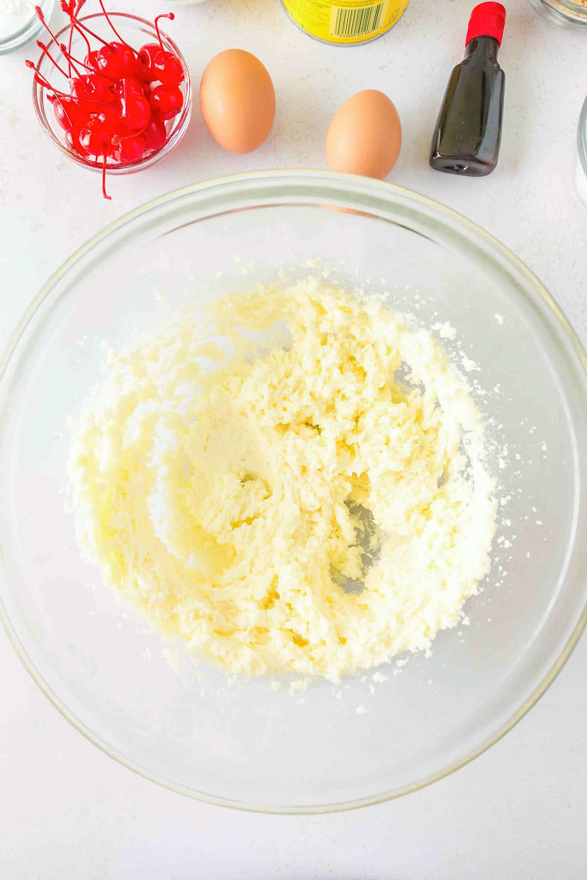A glass bowl with creamed butter and sugar sits on a counter alongside eggs, maraschino cherries, vanilla extract, and other baking ingredientsβeverything you need to start making delicious Pineapple Upside Down Sugar Cookies.