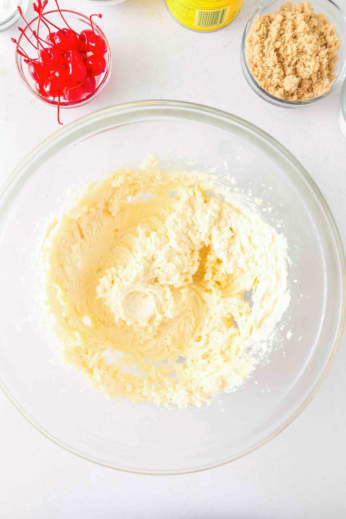 A glass bowl filled with creamed butter and sugar sits on a white counter, surrounded by bowls of brown sugar and maraschino cherriesβessential ingredients for baking delectable Pineapple Upside Down Sugar Cookies.