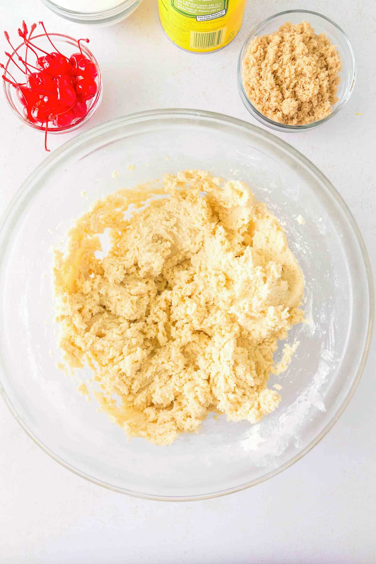 A glass bowl of cookie dough mixture sits on a white surface next to bowls of maraschino cherries, brown sugar, and a can of pineappleβperfect ingredients for making Pineapple Upside Down Sugar Cookies.