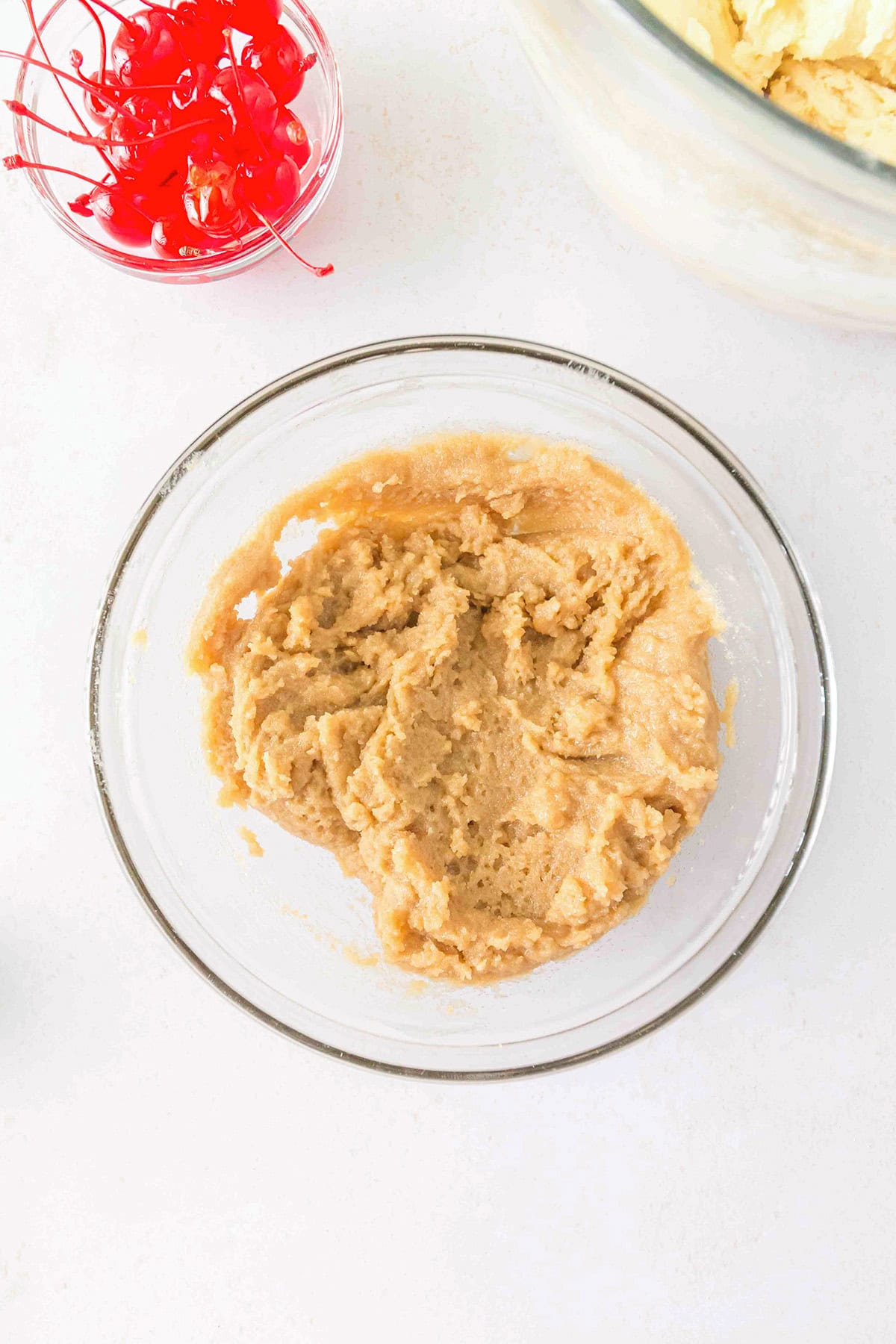 A glass bowl containing cookie doughβperfect for making Pineapple Upside Down Sugar Cookiesβsits on a white surface, with a small bowl of maraschino cherries nearby.