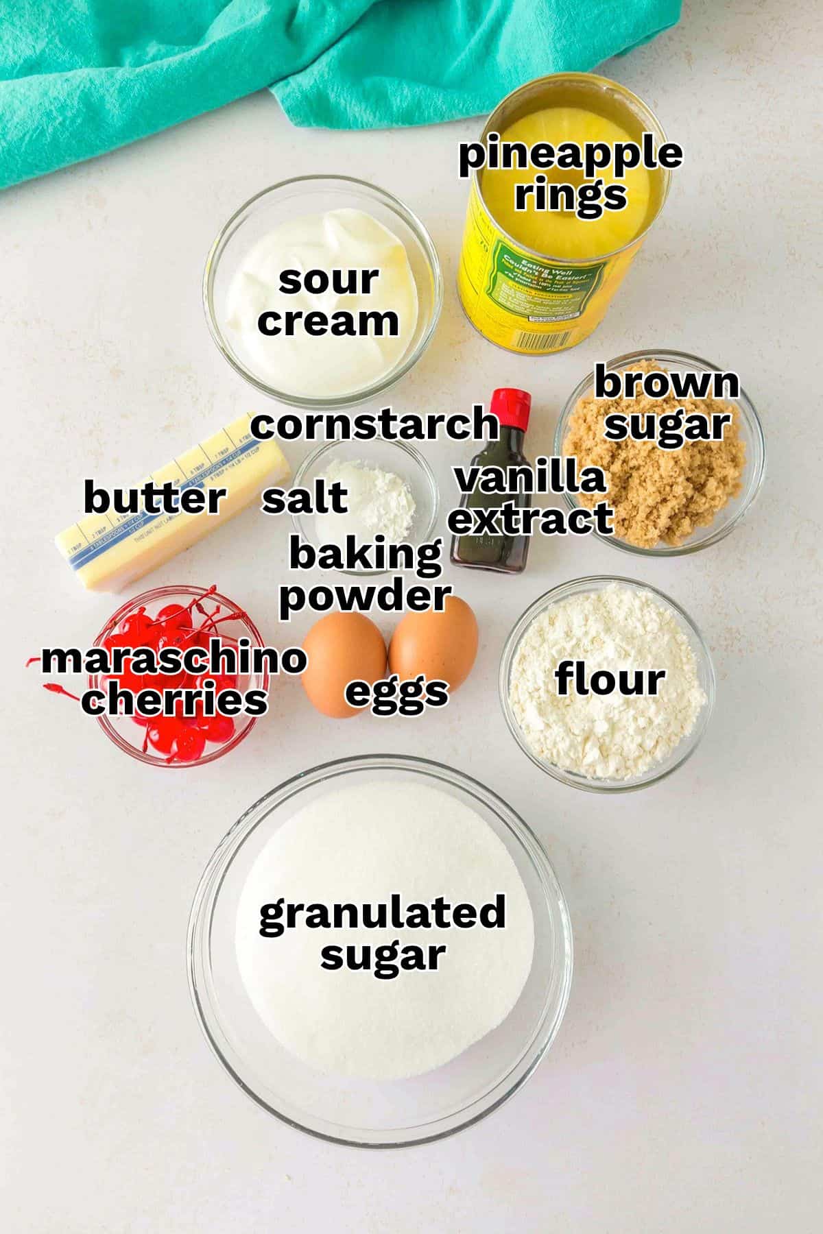 Top-down view of ingredients in bowls and containers labeled: sour cream, pineapple rings, brown sugar, vanilla extract, flour, eggs, granulated sugar, maraschino cherries, butter, salt, cornstarch, and baking powderβall the essentials for making delicious Pineapple Upside Down Sugar Cookies.