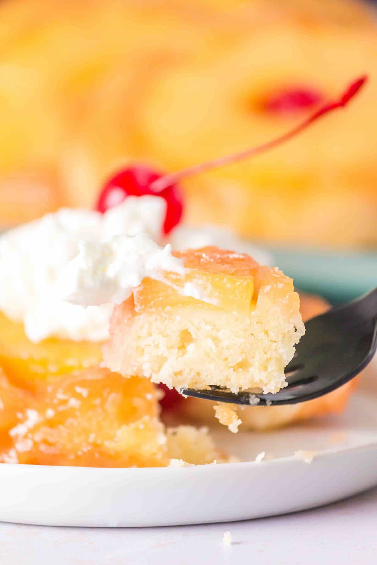 A fork holds a bite of pineapple upside-down cake topped with whipped cream and a cherry, with the rest of the cake on a white plate in the background, reminiscent of the sweet flavors found in Pineapple Upside Down Sugar Cookies.