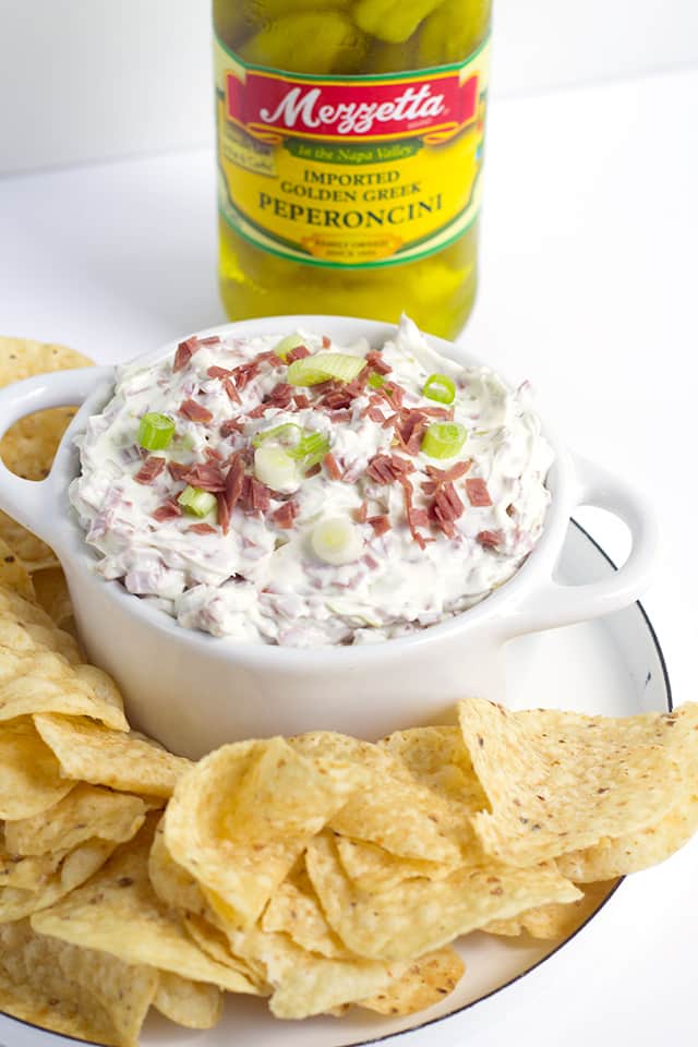 Chipped Beef Dip - This chipped beef dip recipe makes an amazing appetizer for any party. It has dried beef, green onions, and peperoncini in it. It goes great with veggies and bread! This is an easy chip beef recipe that uses dried beef. #appetizer #diprecipe #gamedayrecipes #gamedayfood #partyfood platter of tortilla chips and a bowl of Chipped Beef Dip with a jar of Mezzetta brand Peperoncini behind it