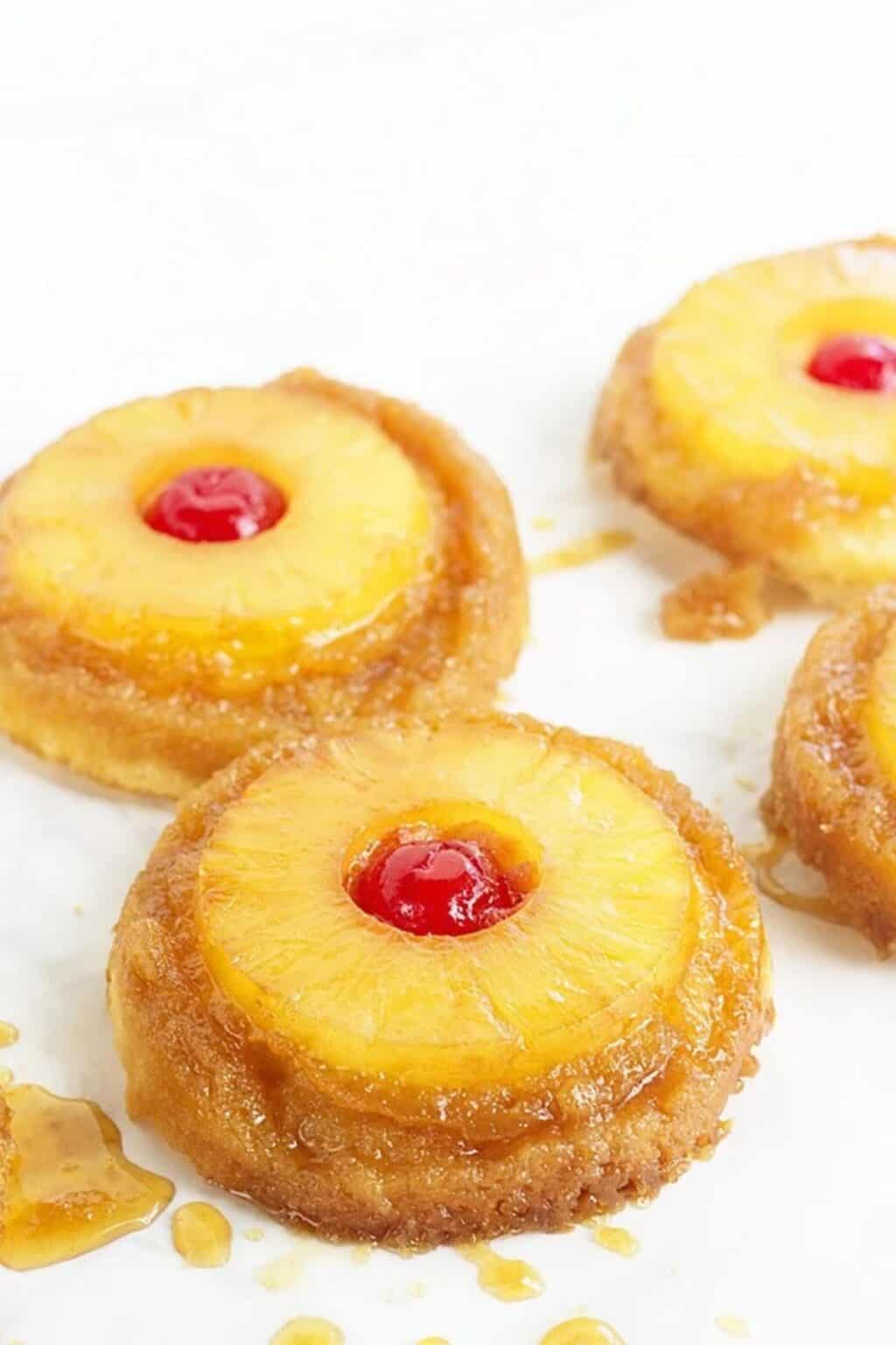 Four mini pineapple upside-down cakes, reminiscent of Pineapple Upside Down Sugar Cookies, topped with pineapple rings and maraschino cherries, arranged on a white surface with some syrup visible.