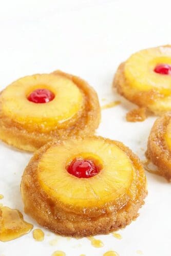 Four mini pineapple upside-down cakes, reminiscent of Pineapple Upside Down Sugar Cookies, topped with pineapple rings and maraschino cherries, arranged on a white surface with some syrup visible.