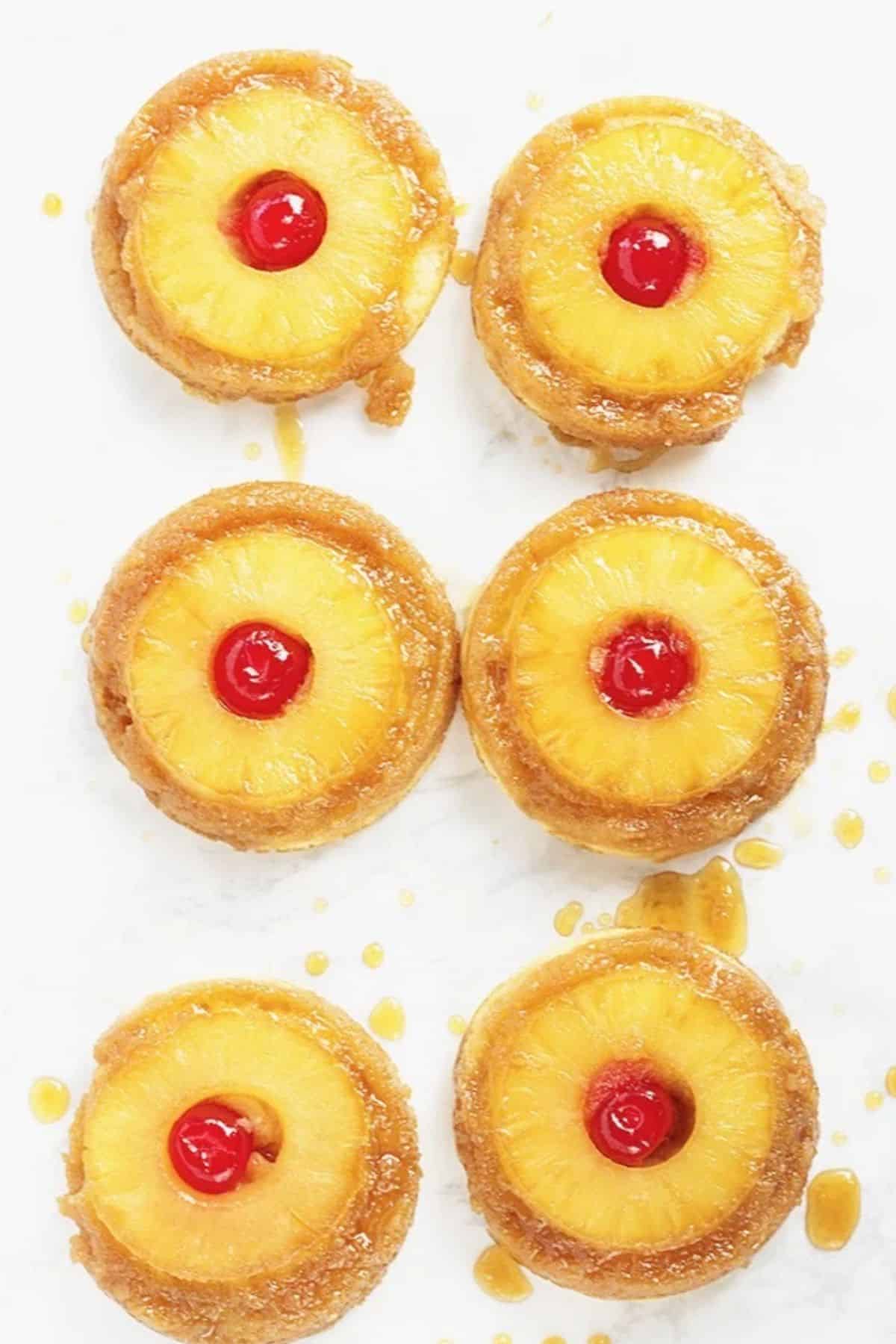 Pineapple Upside Down Sugar Cookies - it's pineapple upside-down cake in cookie form because everyone loves cookies! Soft sugar cookie bottoms with a ring of pineapple and cherry center all covered in a brown sugar glaze! Pineapple upside down sugar cookies are the perfect summer treat and after that tasty pineapple and cherry are gone, you still have a soft, fluffy sugar cookie! These are easy to pick up and eat, but can leave you slightly sticking from all of that buttery brown sugar glaze. #cookies #pineapple #dessertfoodrecipes #cookiedoughandovenmitt Six pineapple upside-down mini cakes topped with pineapple rings and red cherries, arranged on a white surface with caramel sauce dripsβthese treats are reminiscent of Pineapple Upside Down Sugar Cookies in both flavor and style.