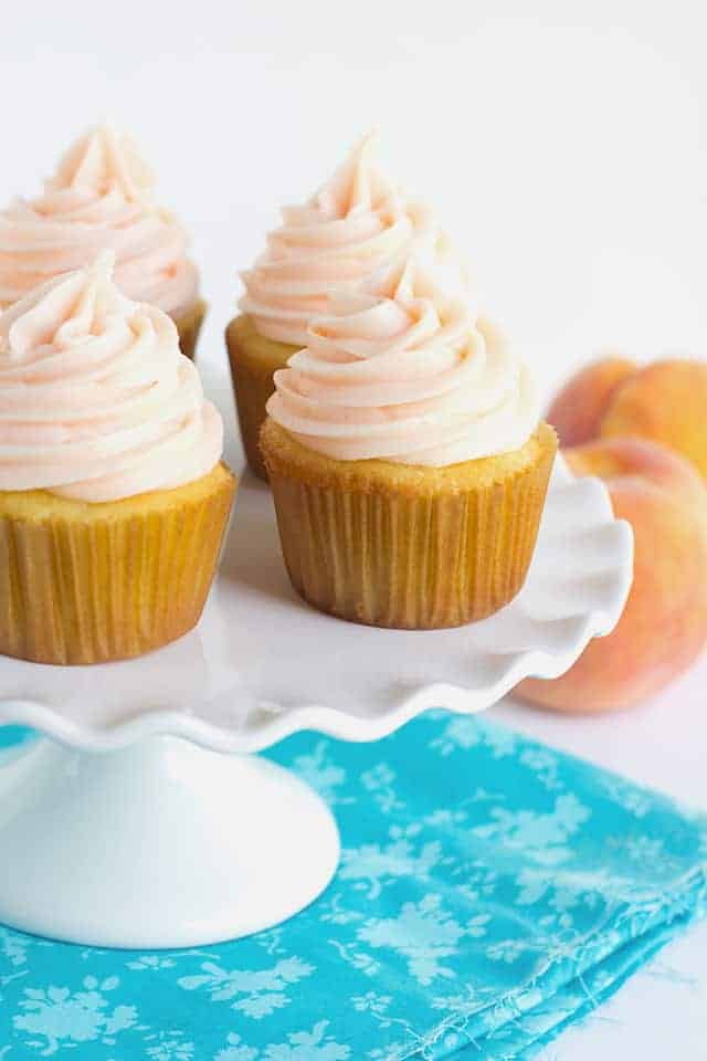 Yellow Cupcakes with Peach Cream Cheese Frosting - These yellow cupcakes with peach cream cheese frosting need to happen at every party. The yellow cake is so light and fluffy and the peach cream cheese frosting has a nice pop of flavor! Did I mention that peach colored frosting? #cookiedoughandovenmitt #cupcakes #cupcakerecipe #yellowcake #vanillacake #vanillacakerecipe #dessertfoodrecipes yellow cupcakes on a white cake plate with blue fabric
