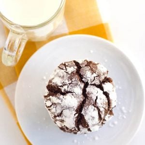Flat lay photo of a chocolate crinkle cookie on with a yellow and white checkered linen and a cup of milk
