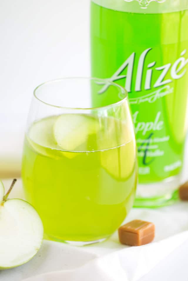 Caramel Apple Cocktail - how fun is a bright green drink that's caramel apple flavored?! This is a sweet cocktail that packs a punch from the liqueur. Garnish with a couple of apple slices and serve at your next party!