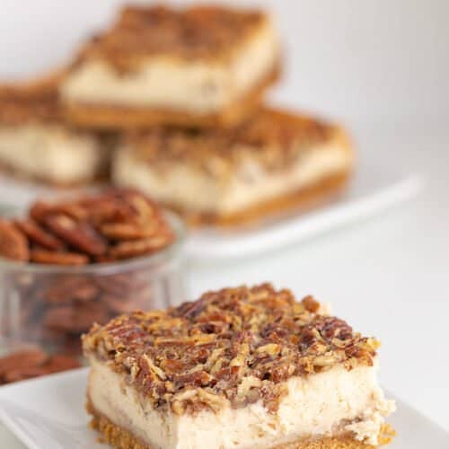 cheesecake bar on a white square plate with pecans and cheesecake bars stacked behind it