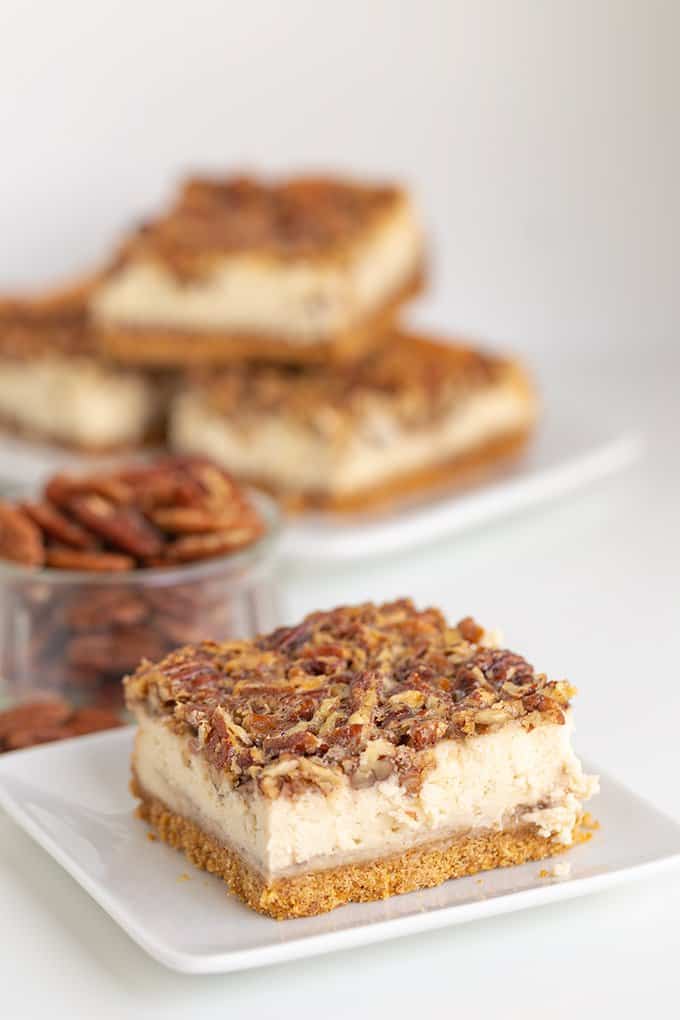 cheesecake bar on a white square plate with pecans and cheesecake bars stacked behind it