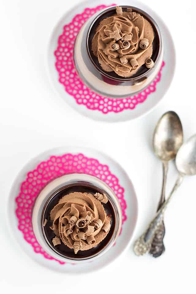 Standing over top of two of the two trifle glasses filled with layers of chocolate trifle on a pink doily and white saucer