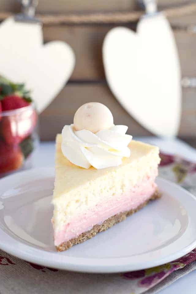 a slice of strawberries and cream cheesecake sitting on a plate with a floral napkin underneath and strawberries in a glass bowl behind it