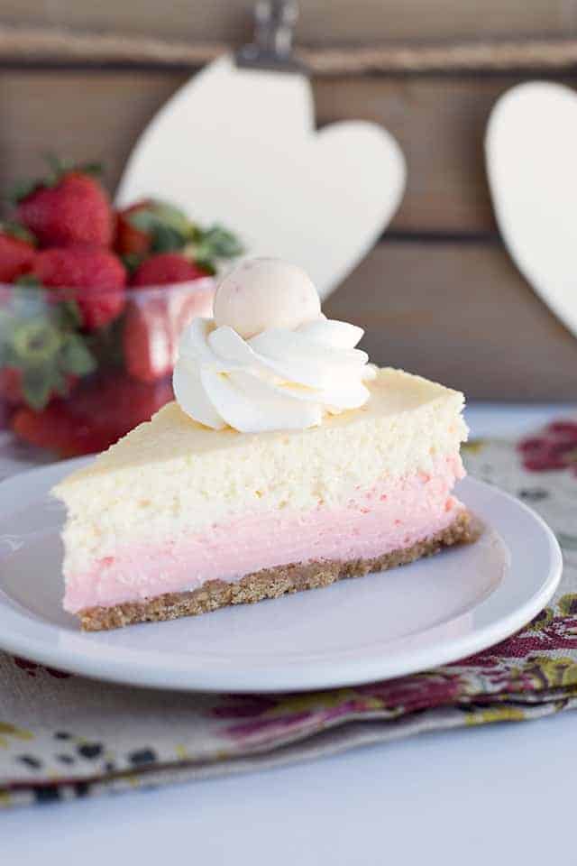 Strawberries and Cream Cheesecake - This cheesecake is packed full of flavor! There's a cookie crust, pink strawberry cheesecake layer, vanilla cheesecake layer, strawberries and cream truffles baked inside the cheesecake and topped with whipped cream and more strawberries and cream truffles! This is the perfect Valentine's Day cheesecake! #cookiedoughandovenmitt #cheesecake #cheesecakerecipes #dessertrecipes #strawberries a slice of strawberries and cream cheesecake sitting on a plate with a floral napkin underneath and strawberries in a glass bowl behind it