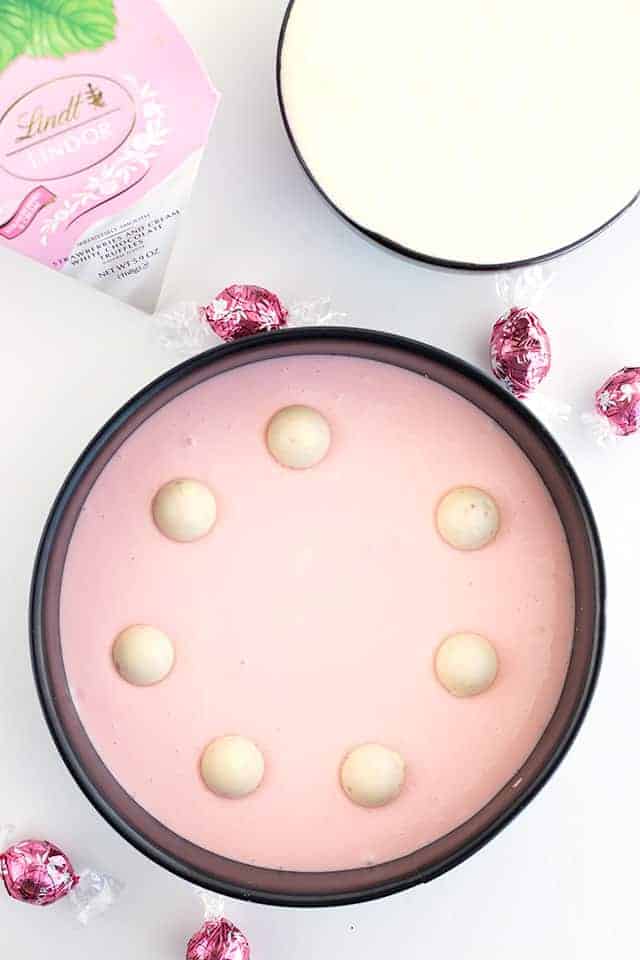 Pink cheesecake batter is in a springform pan with strawberries and cream truffles added in.