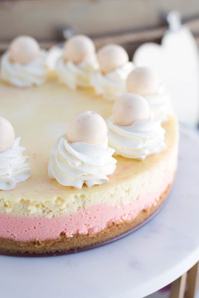 Strawberries and Cream Cheesecake - This cheesecake is packed full of flavor! There's a cookie crust, pink strawberry cheesecake layer, vanilla cheesecake layer, strawberries and cream truffles baked inside the cheesecake and topped with whipped cream and more strawberries and cream truffles! This is the perfect Valentine's Day cheesecake! #cookiedoughandovenmitt #cheesecake #cheesecakerecipes #dessertrecipes #strawberries Full strawberries and cream cheesecake sitting on a marble cake stand