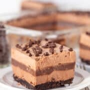 A decadent slice of chocolate layer dessert sits on a plate, featuring a luscious whipped cream top and rich chocolate drizzle over a crisp crust base. In the background, more tempting slices await.