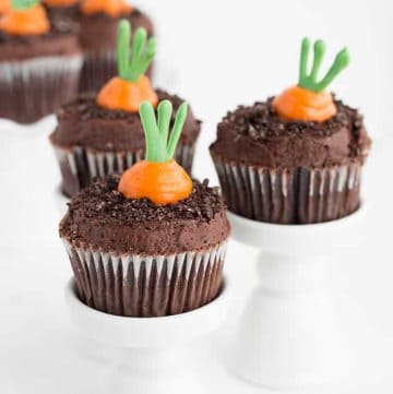 Three carrot patch cupcakes sitting on individual cupcake stands with carrot patch cupcakes on a ruffled white cake stand in the background