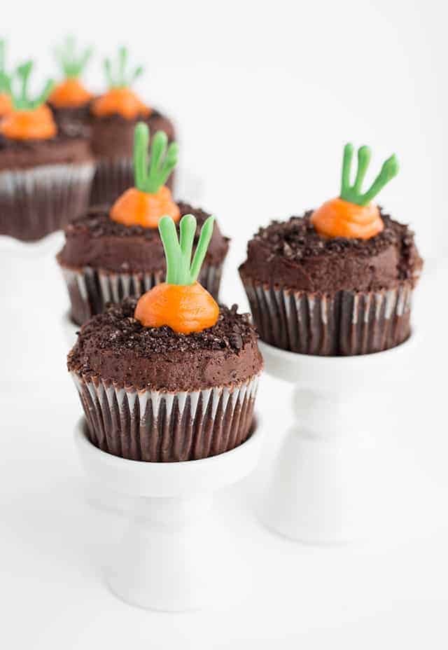 Three carrot patch cupcakes sitting on individual cupcake stands with carrot patch cupcakes on a ruffled white cake stand in the background