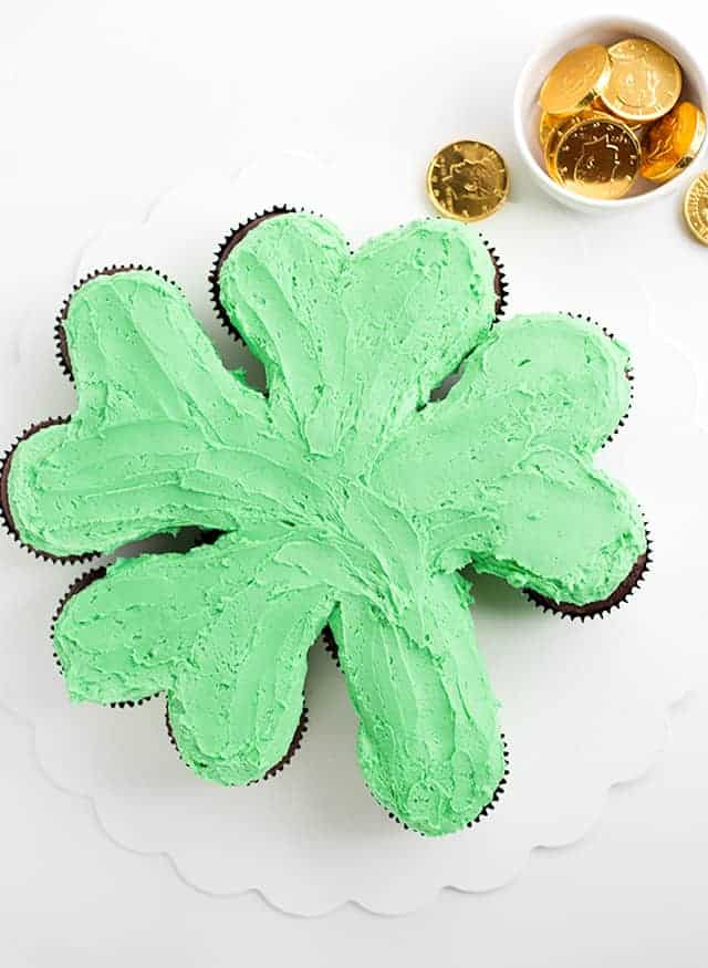 St. Patrick's Day Shamrock Cupcake Cake - The cupcakes are made from a doctored up cake mix and have a great chocolate mint flavor! They're topped with a gorgeous green minty frosting and rainbow sprinkles! #cookiedoughandovenmitt #StPatricksDay #cupcakecake #cakes #dessertrecipes #dessertfoodrecipe Overhead shot of green shamrock cupcake cake on a white cake board.