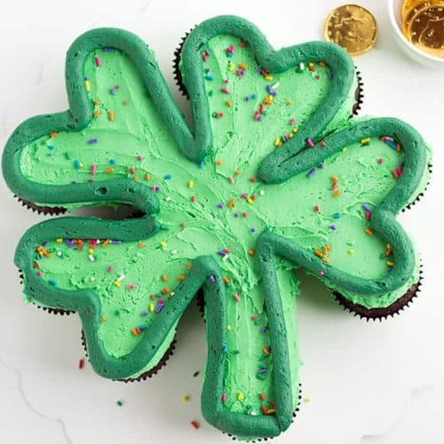 A green shamrock cupcake cake with a dark green outline and rainbow sprinkles on a white cake board with gold chocolate coins scattered.