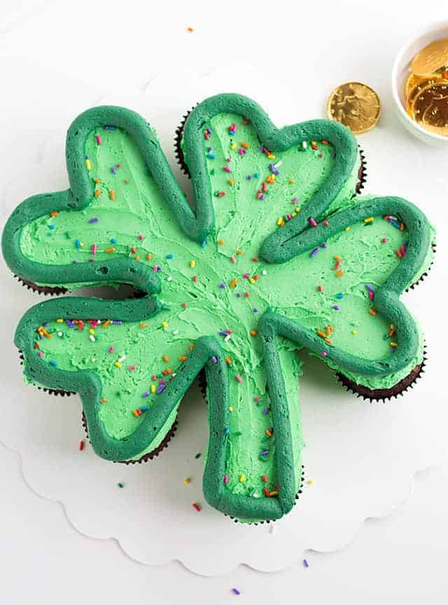 A green shamrock cupcake cake with a dark green outline and rainbow sprinkles on a white cake board with gold chocolate coins scattered.