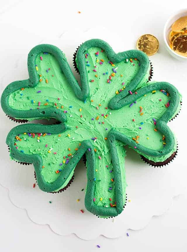 St. Patrick's Day Shamrock Cupcake Cake - The cupcakes are made from a doctored up cake mix and have a great chocolate mint flavor! They're topped with a gorgeous green minty frosting and rainbow sprinkles! #cookiedoughandovenmitt #StPatricksDay #cupcakecake #cakes #dessertrecipes #dessertfoodrecipe A green shamrock cupcake cake on a white cake board with gold chocolate coins scattered.