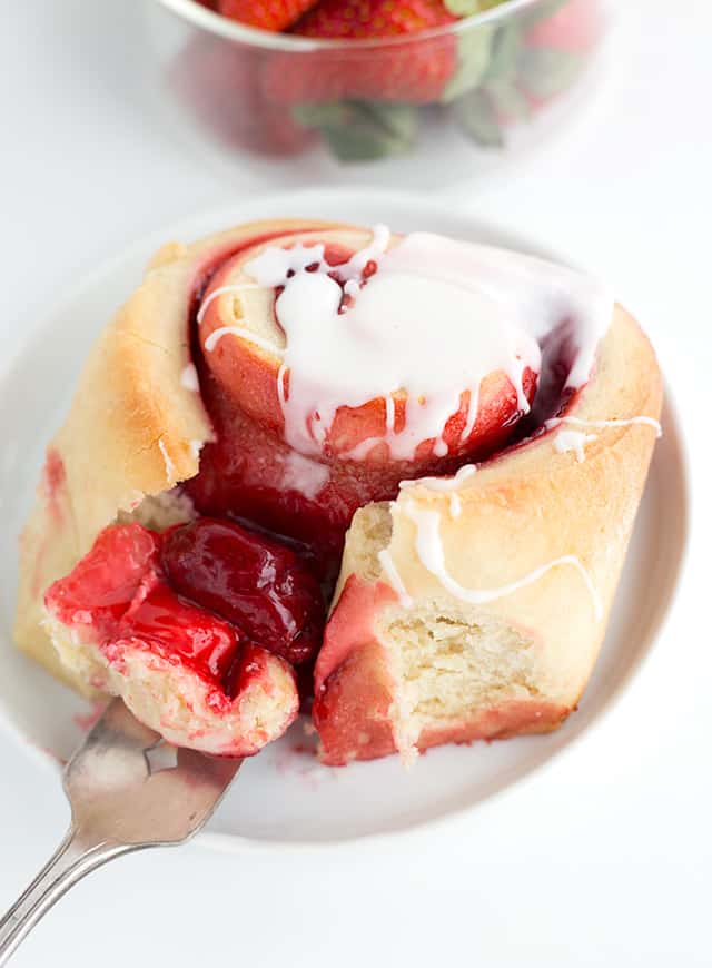 A fork full of the strawberry sweet rolls is showing. It's a single sweet roll on a white plate with glaze on top.