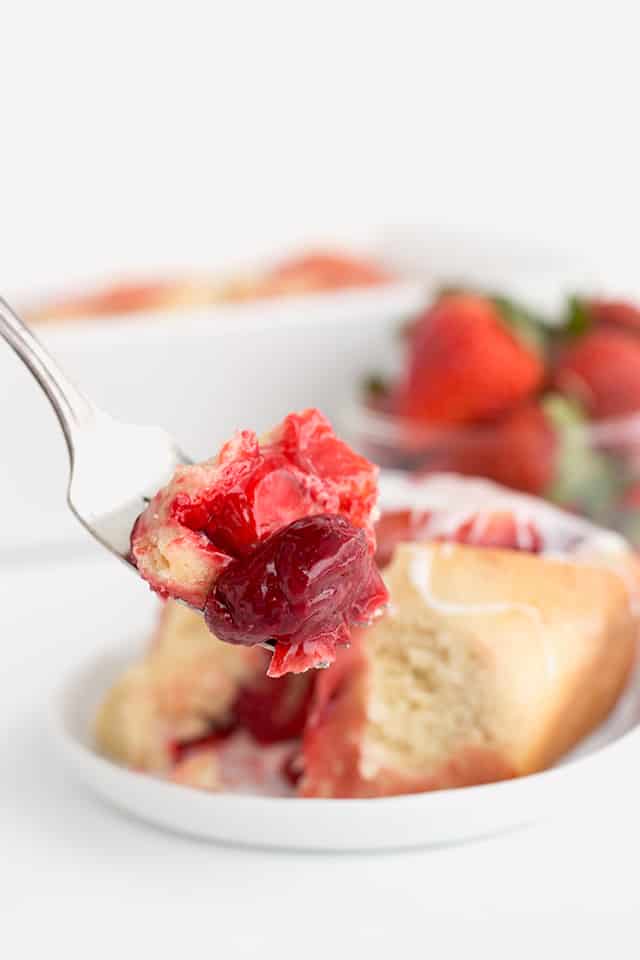 A fork full of the strawberry sweet rolls with a strawberry is in focus. In the background is a sweet roll on a white plate, strawberries in a bowl, and the white pan.