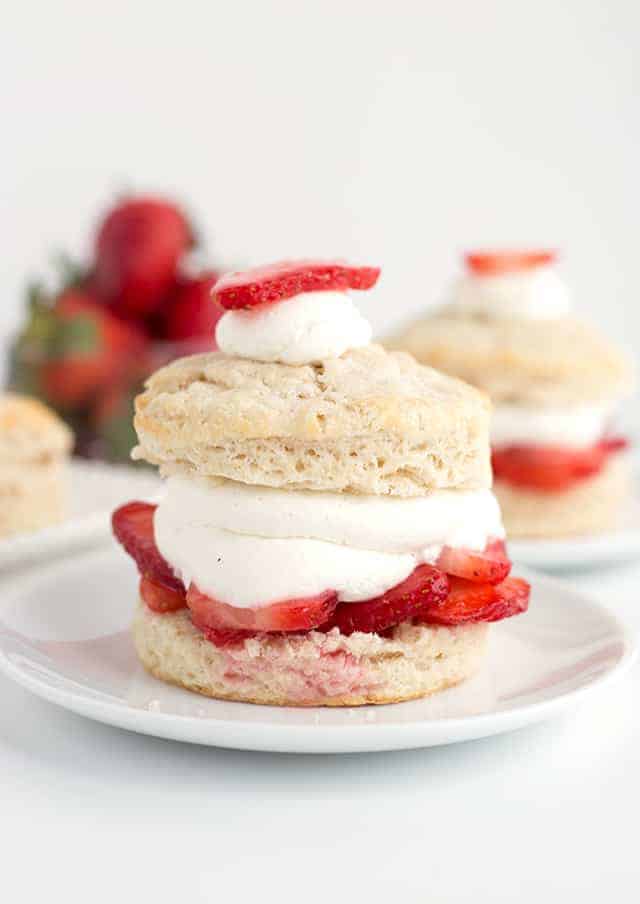 Close up of homemade strawberry shortcake on a small white plate with strawberries and another strawberry shortcake in the back.