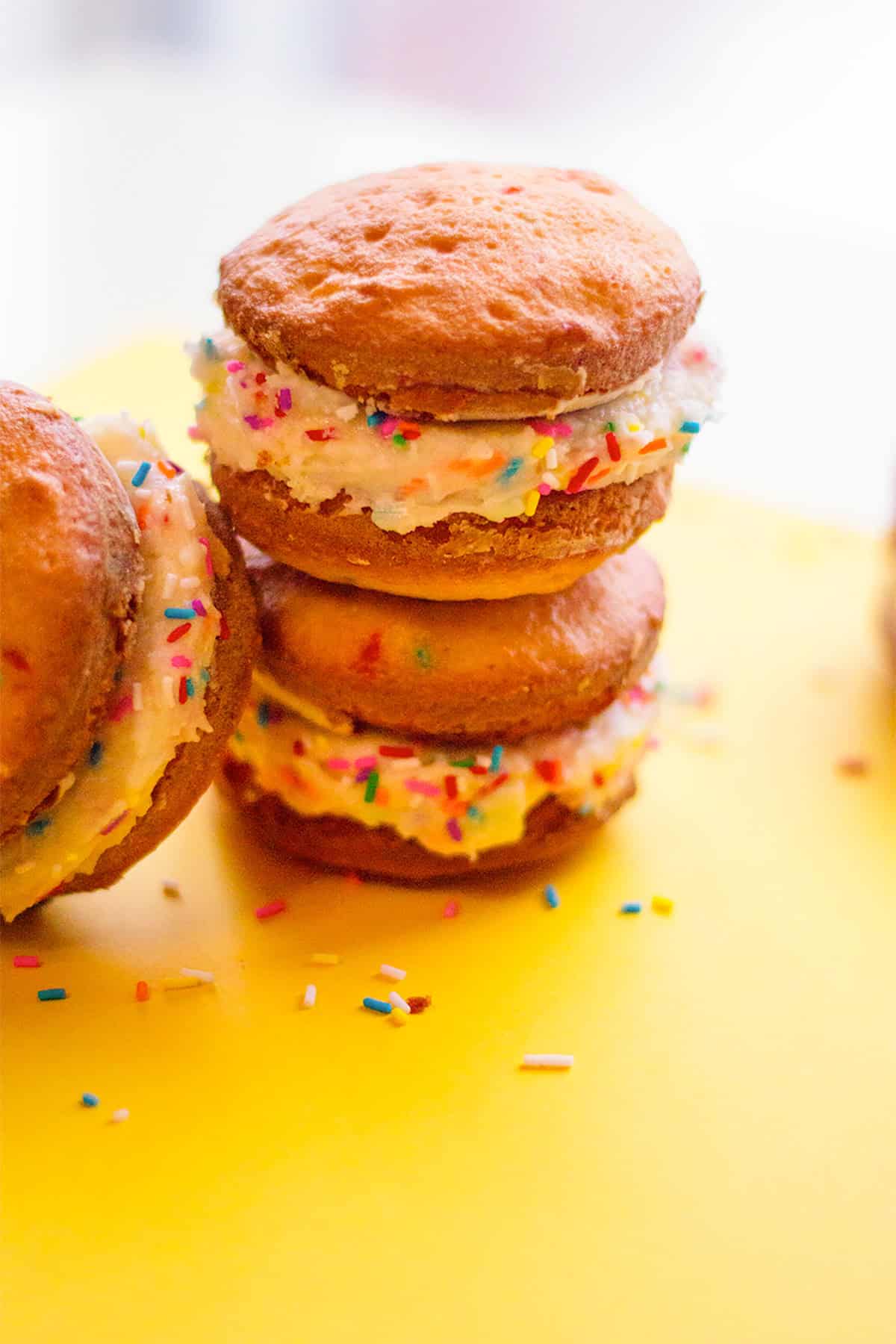 Two cake mix whoopie pies with cream filling and colorful sprinkles are stacked on a yellow surface, with loose sprinkles scattered nearby.