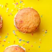 Cake mix whoopie pies with white cream filling and colorful sprinkles are arranged on a bright yellow surface, with extra sprinkles scattered around.
