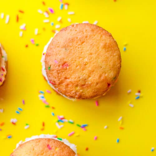 Cake mix whoopie pies with white cream filling and colorful sprinkles are arranged on a bright yellow surface, with extra sprinkles scattered around.