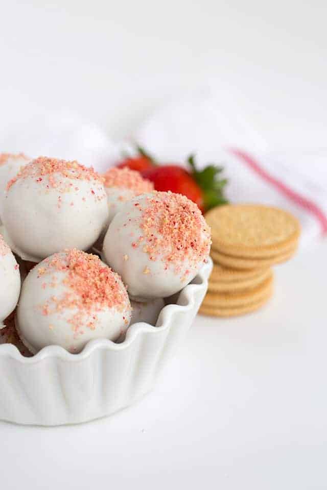 Half of the white bowl of strawberry shortcake truffles in the picture with a white and red hand towel and ingredients behind them