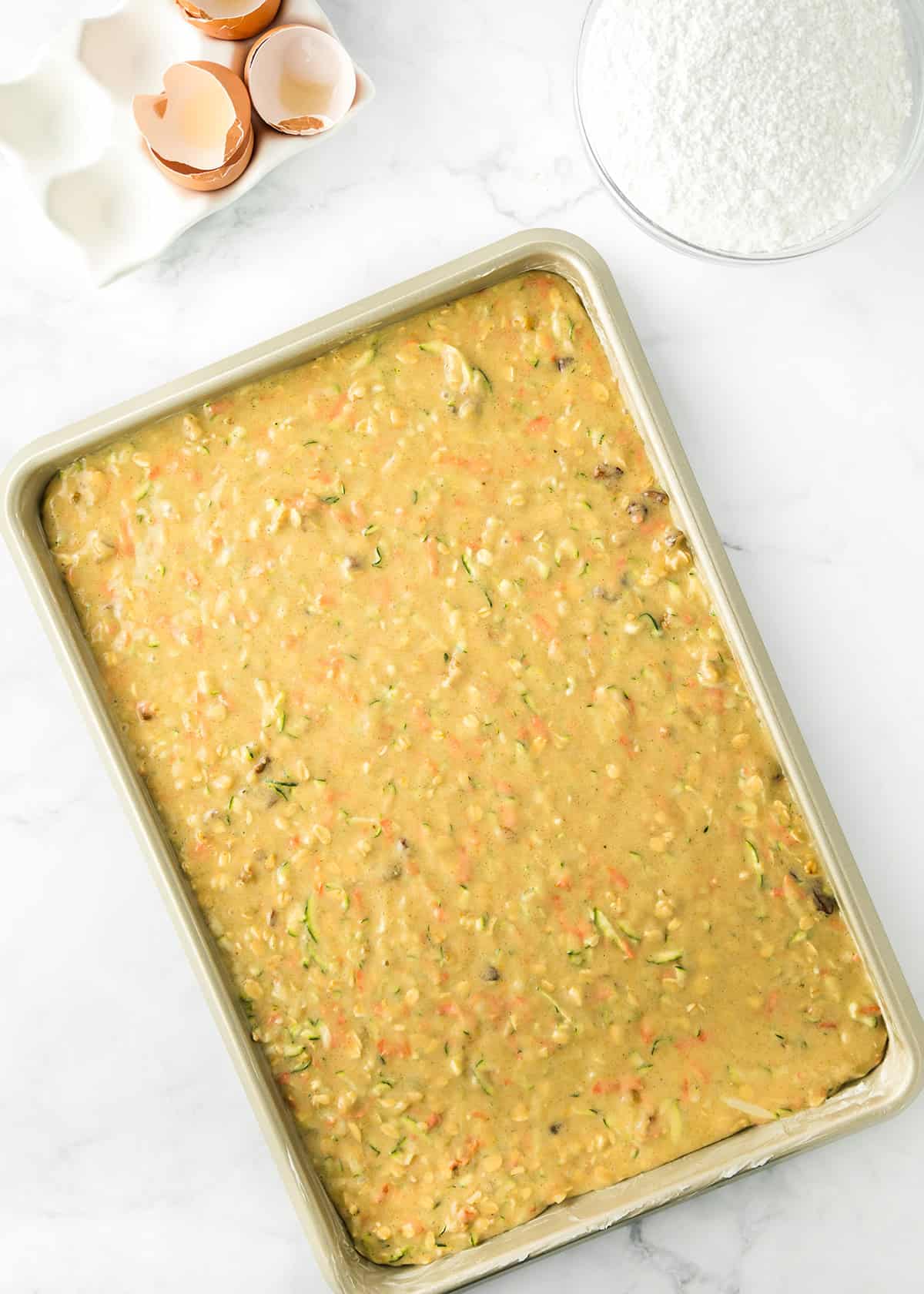 Rectangular baking pan filled with uncooked zucchini bars batter sits on a white surface beside cracked eggshells and a bowl of shredded coconut.