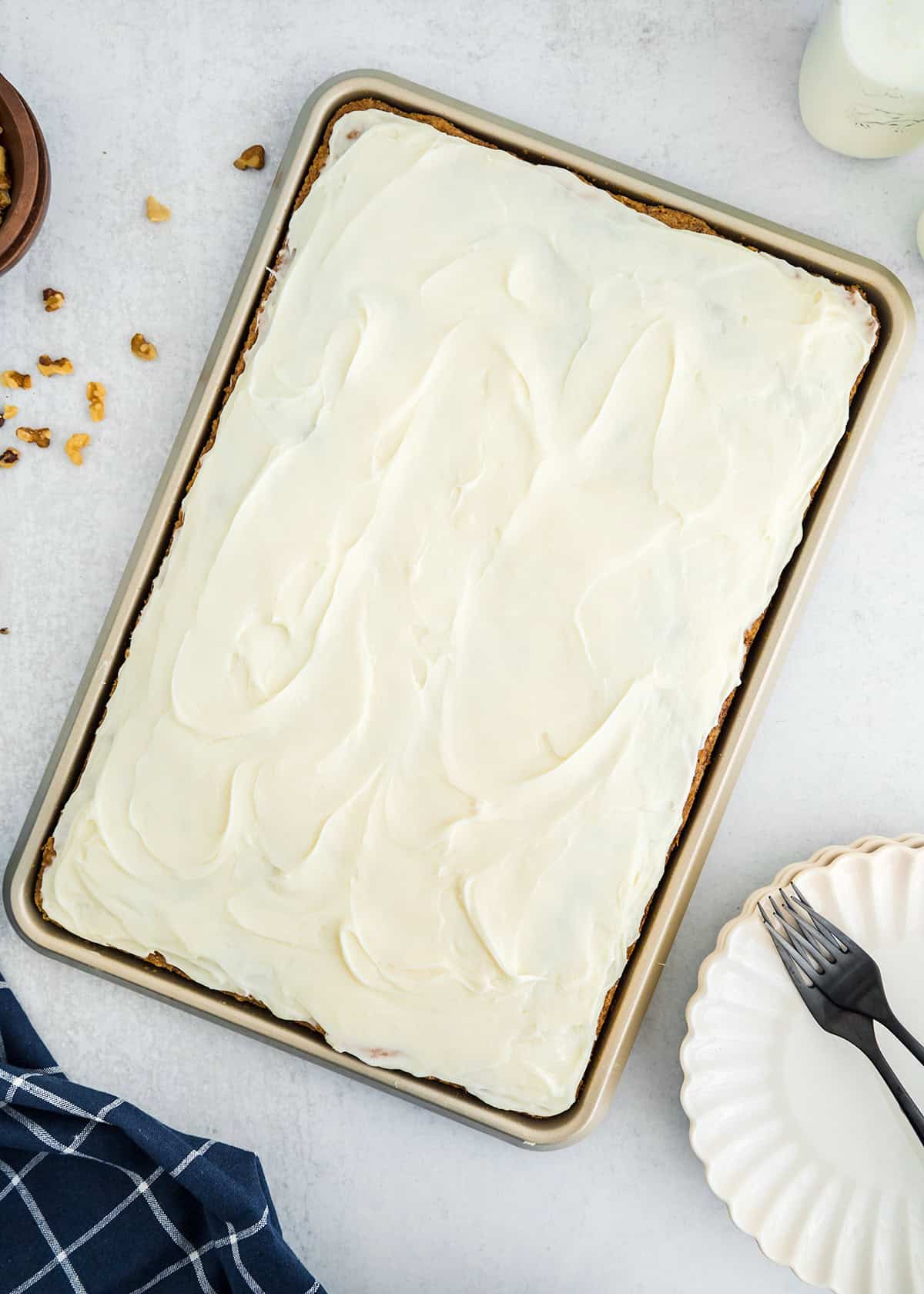 A rectangular sheet cake with white frosting on a baking tray, surrounded by a fork, plate, walnuts, and a blue plaid cloth—perfectly capturing the cozy charm of homemade zucchini bars.