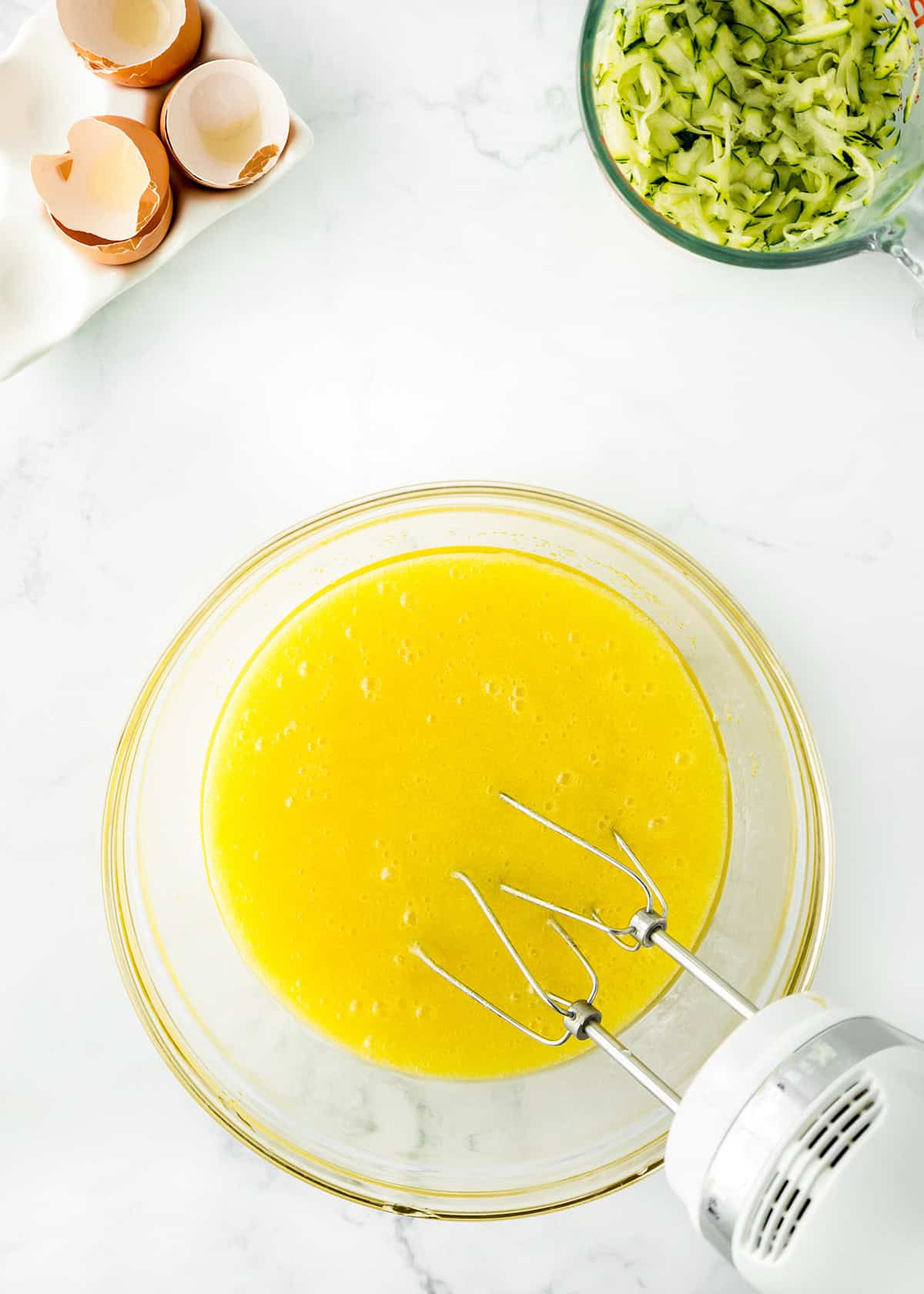 A glass bowl of beaten eggs is being mixed with a hand mixer; empty eggshells and a bowl of shredded zucchini are nearby on a white countertop, ready for making delicious zucchini bars.