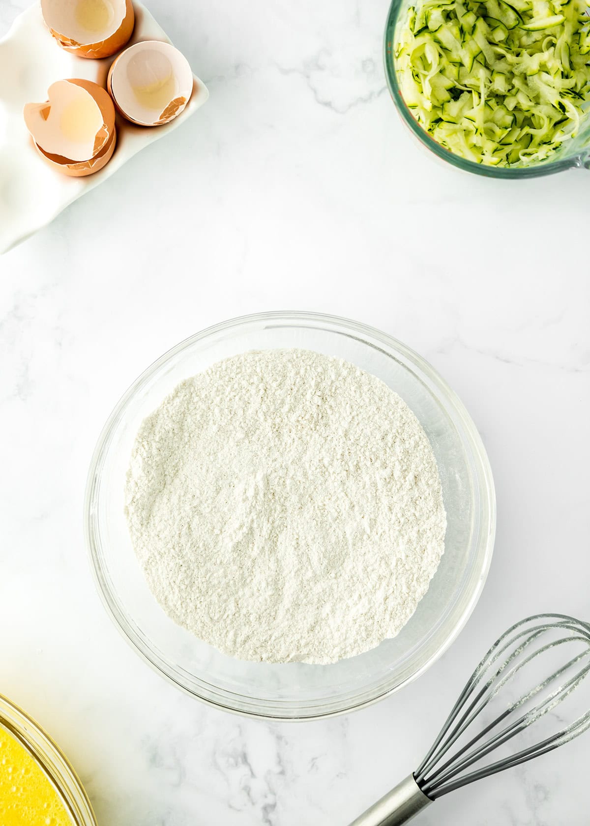 A glass bowl of flour, a whisk, cracked eggs in a carton, and a bowl of shredded zucchini are arranged on a white marble surface—everything you need to start making delicious zucchini bars.