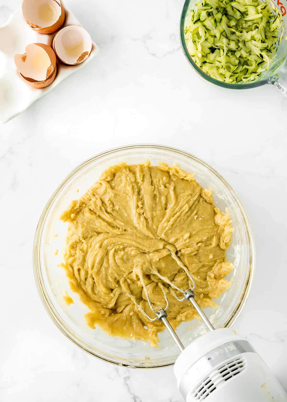 A mixing bowl with batter and a hand mixer, a bowl of shredded zucchini for delicious zucchini bars, and cracked eggshells on a marble countertop.