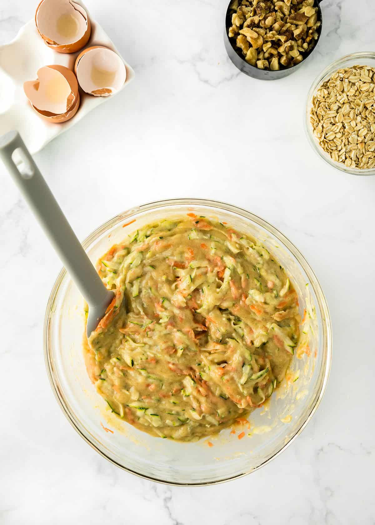 A glass bowl of mixed zucchini bars batter with a spatula, surrounded by eggshells, walnuts, and oats on a white countertop.