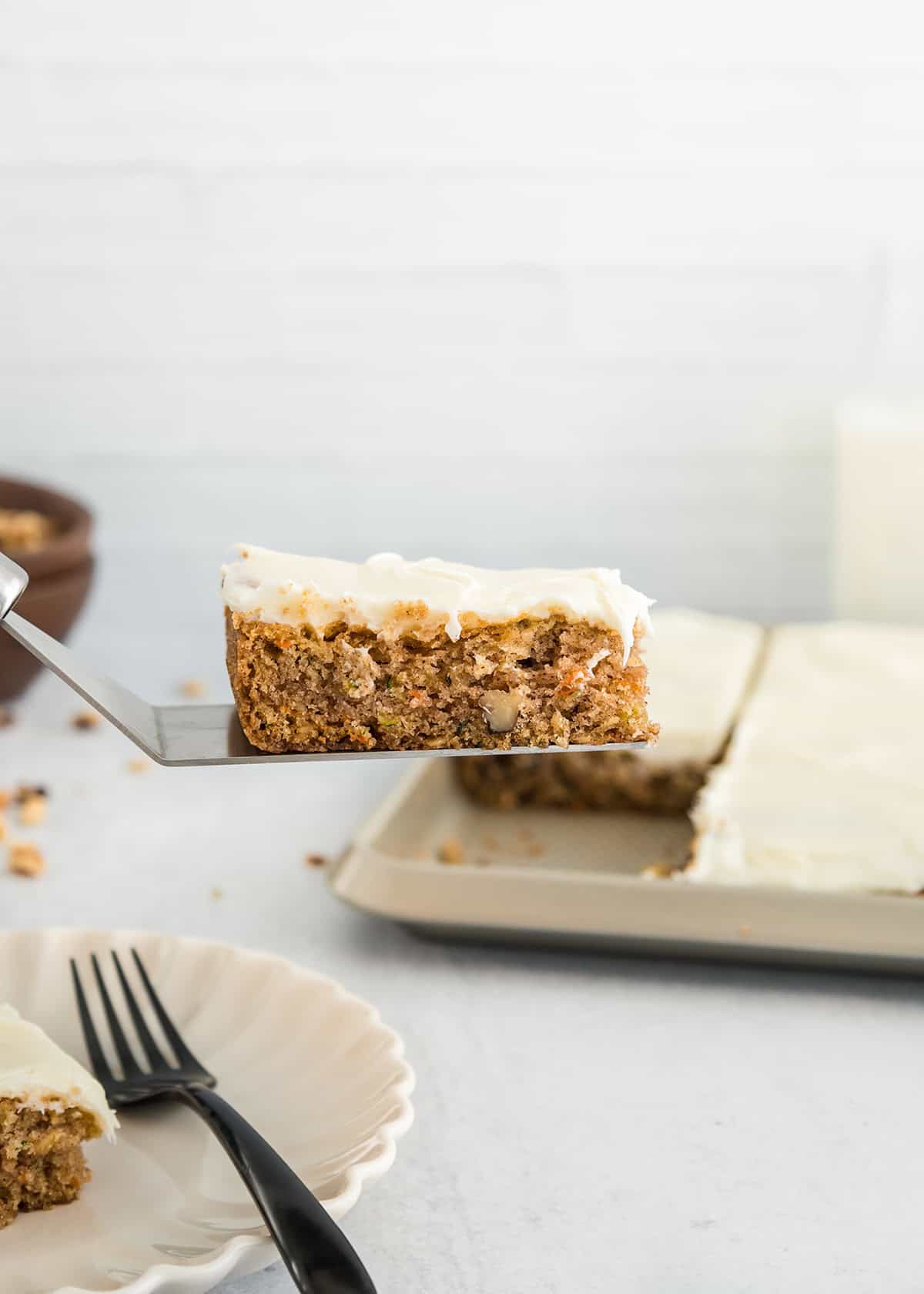 A spatula holds a slice of frosted cake with visible nuts, above a plate and a baking pan containing more zucchini bars in the background.