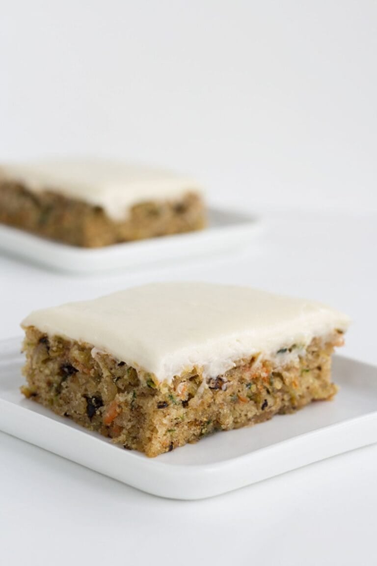 Two square pieces of frosted zucchini carrot cake, reminiscent of classic zucchini bars, sit on white plates against a plain white background.