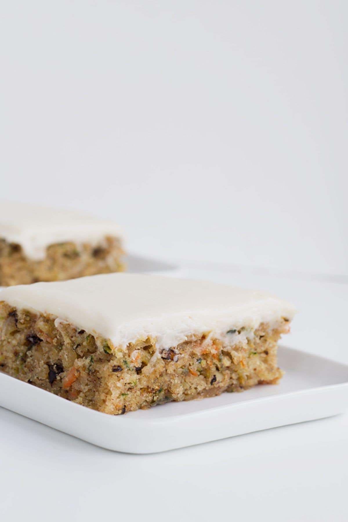 A square slice of frosted carrot cake with visible nuts and carrot pieces sits on a white rectangular plate, reminiscent of homemade zucchini bars in its moist texture and inviting appearance.