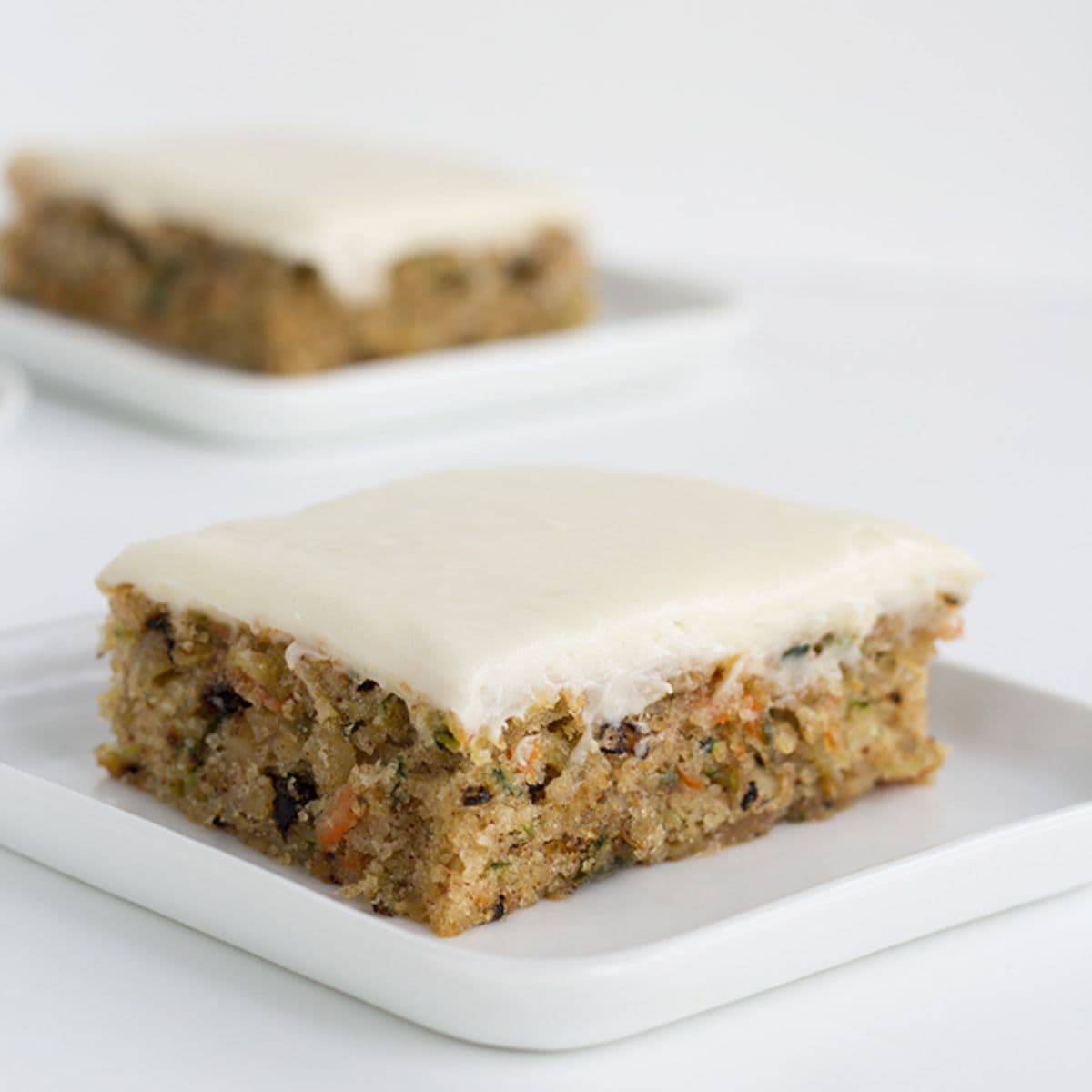 A square piece of zucchini cake with cream cheese frosting sits on a white plate, reminiscent of classic zucchini bars, with another piece blurred in the background.