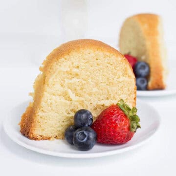 A slice of vanilla pound cake on a white plate with strawberries and blueberries, with another plate of cake in the background.