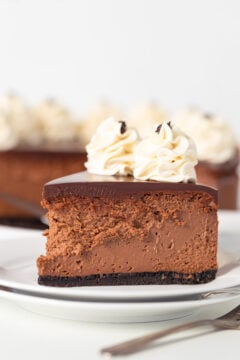 A slice of chocolate mousse Baileys cheesecake features a dark chocolate layer and is adorned with two swirls of whipped cream. It rests elegantly on a white plate, accompanied by forks waiting to indulge.