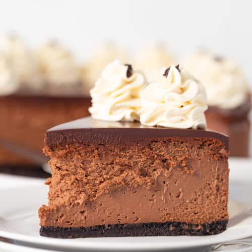 A slice of chocolate mousse Baileys cheesecake features a dark chocolate layer and is adorned with two swirls of whipped cream. It rests elegantly on a white plate, accompanied by forks waiting to indulge.
