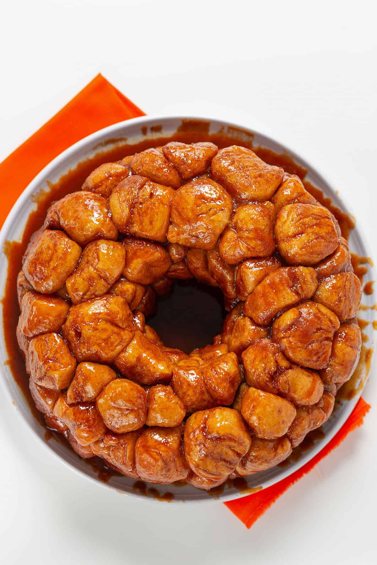A freshly baked monkey bread, crafted from frozen bread dough, sits in a ring shape on a white plate with an orange napkin underneath.
