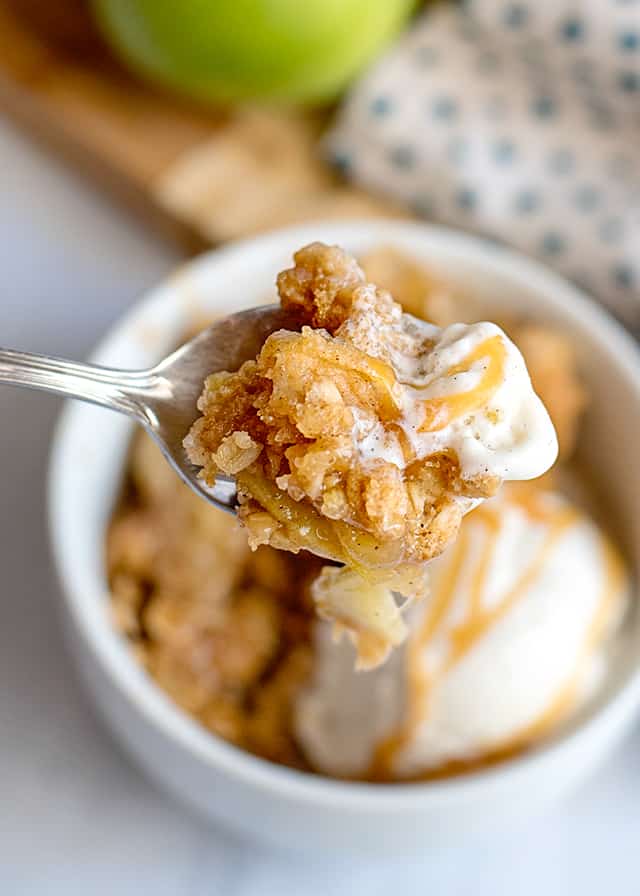 Spoonful of vanilla ice cream and caramel apple crisp with white bowl in background