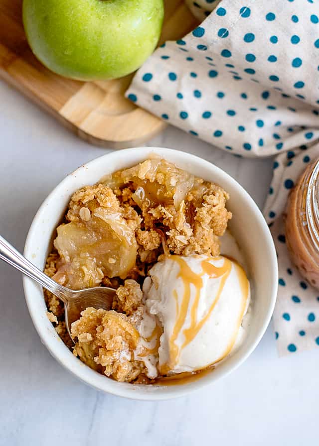 spoon in white bowl with caramel apple crisp, vanilla ice cream, and caramel sauce